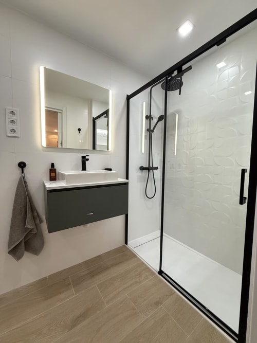Baño moderno en apartamento de Fuengirola con accesorios negros, espejo iluminado, ducha abierta y piso efecto madera
