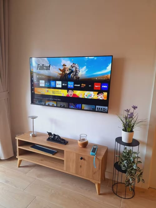 Sala de estar de apartamento moderno con TV montado en la pared mostrando aplicaciones de streaming, mueble de madera para TV, plantas en macetas y elegante