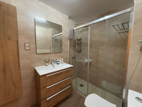 Baño moderno con tocador de madera, cabina de ducha de cristal y paredes alicatadas en apartamento de Fuengirola