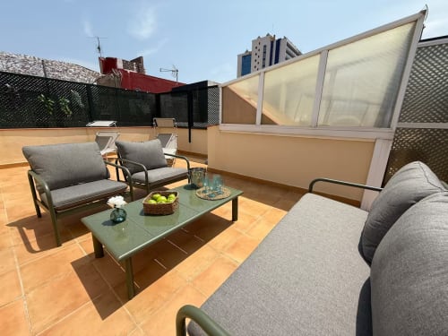Terraza soleada en la azotea con asientos grises, mesa verde y vistas de la ciudad en apartamento de Fuengirola