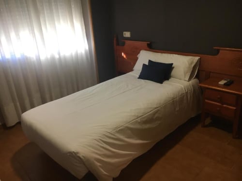 Habitación individual en Hostal Imaginatelo con cama de madera, sábanas blancas, almohada oscura, mesita de noche y ventana con cortina translúcida