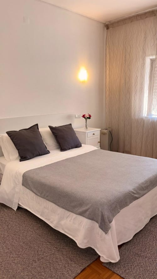 Quarto de casal elegante na Porta 80 House Braga com roupa de cama cinzenta, almofadas escuras e iluminação acolhedora de arandela