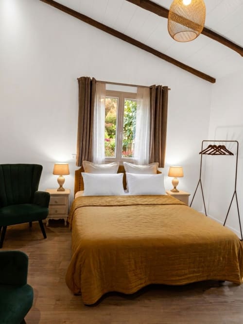Chambre lumineuse à La Bastide Des Muriers avec couvre-lit doré, oreillers blancs, fauteuil vert, poutres en bois et vue sur le jardin