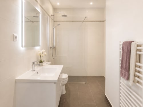 Salle de bain moderne de l'appartement Le FETTOLINA à Cannes avec douche à l'italienne, vasque blanche, porte-serviettes chauffant et éclairage du miroir