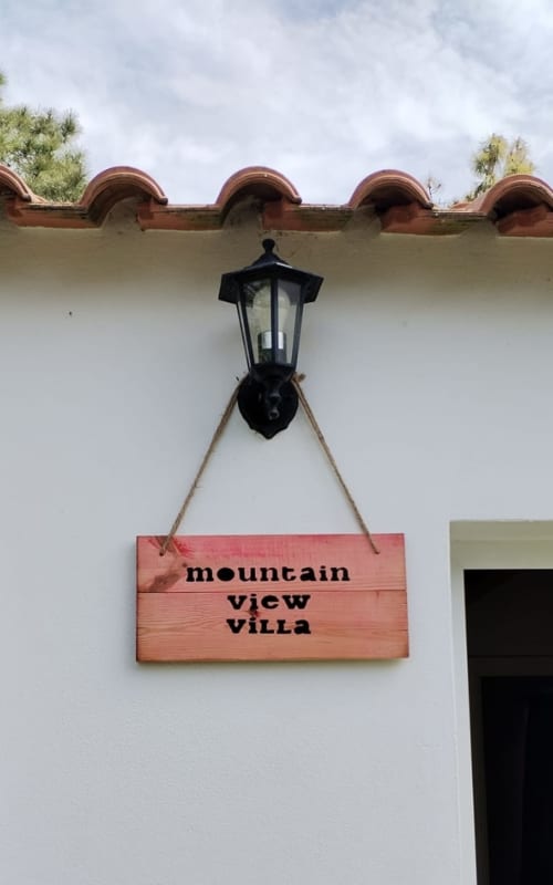 Wooden sign reading 'mountain view villa' hanging from black lamp on white wall with terracotta roof tiles