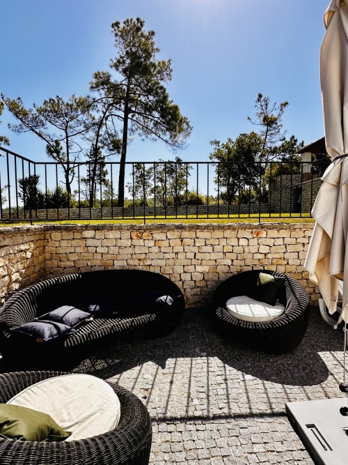 Luxury patio lounge at Quinta Boa Vida cottage in Alcobaça with woven chairs, stone wall, pine trees, and clear blue sky
