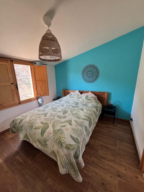 Quarto turquesa brilhante com cama de madeira, roupa de cama com padrão de samambaia, luminária suspensa tecida e vista para o jardim