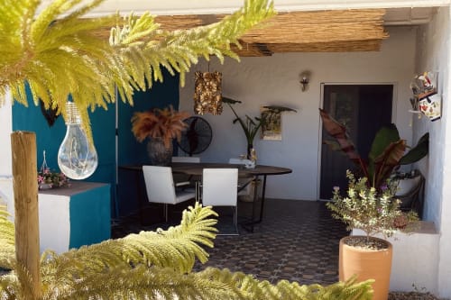 Pátio rústico de cortijo andaluz com frondes de palmeira amarelas, área de refeições, plantas em vasos e parede de destaque azul-petróleo em Huércal-Overa.