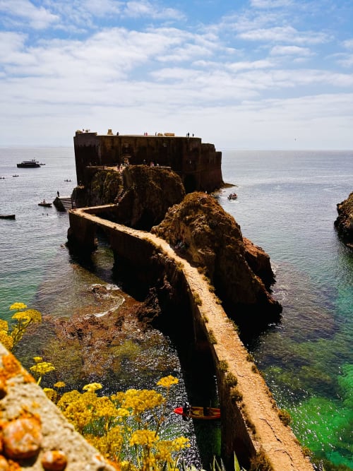 Ancient stone fortress on rocky island off Portuguese coast with yellow wildflowers, clear blue sea, and boats nearby