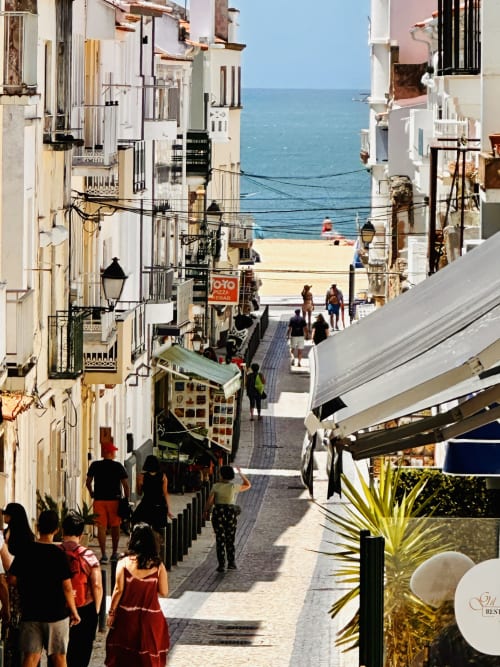 Narrow cobblestone street descending to beach in Alcobaça, Portugal, lined with white buildings and shops, blue sea visible