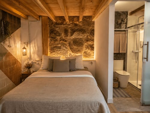 Dormitorio acogedor en la casita Holibai de Baiona con muros de piedra, vigas de madera, iluminación ambiental y baño ensuite