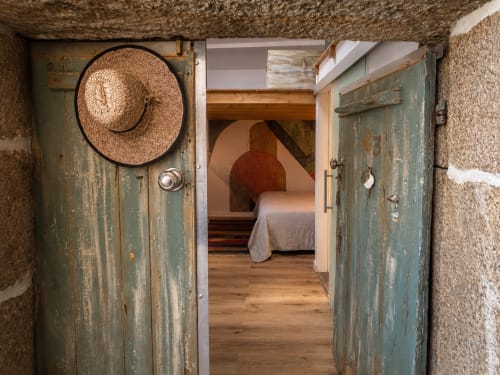 Puertas de madera rústica abiertas al dormitorio de la casita Mirada do Roxo, Baiona, con sombrero de paja y muros de piedra