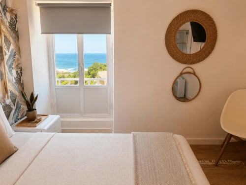 Luminoso dormitorio costero en cabaña de Baiona con vistas al mar, ropa de cama crema, espejos tejidos y ventana con vista al océano azul