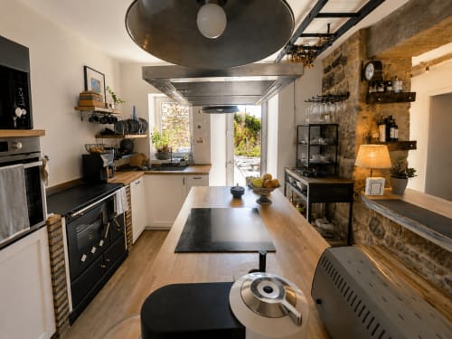 Cocina de cabaña moderna en Baiona con encimeras de madera, electrodomésticos negros, paredes de piedra y vista al jardín a través de puertas de cristal