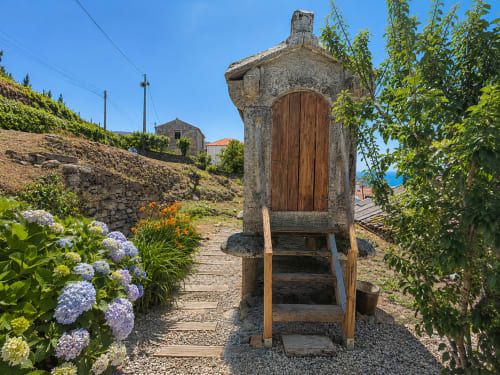 Entrada rústica de casa de piedra con puerta de madera en Holibai, Baiona, rodeada de flores azules y enredaderas verdes