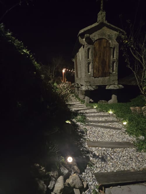 Sendero nocturno hacia casita de piedra en Holibai, Baiona, España, con iluminación de jardín y vista al mar