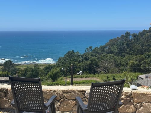 Dos sillas negras en terraza de piedra con vistas al Mediterráneo, vegetación exuberante y costa gallega en la casita Holibai,