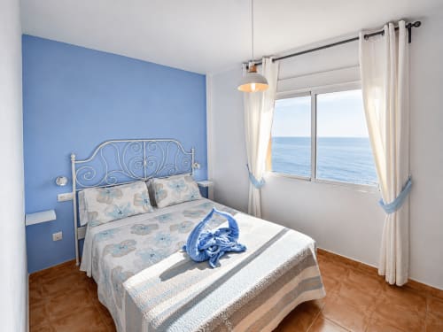 Luminosa habitación con pared azul destacada, cama ornamentada y gran ventanal con vistas al mar en Apartamentos El Calón Playa, Cuevas del