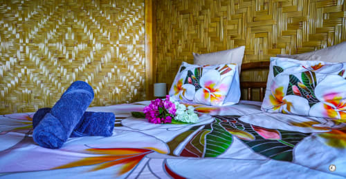 Serviettes bleues roulées et fleurs tropicales sur un lit aux coussins colorés à la villa Aimehome à Teavaro