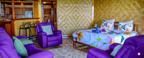 Chambre de la villa Kia Ora avec fauteuils violets, lit aux motifs floraux colorés, murs tressés et équipements modernes à Teavaro