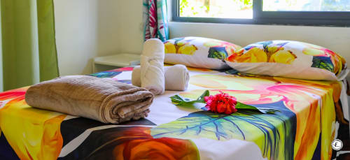 Literie tropicale colorée aux motifs floraux sur un lit de villa à Moorea, avec coussins déccoratifs et fleur rouge fraîche