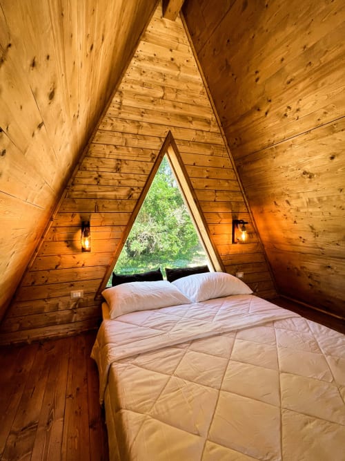 Cozy A-Frame glamping bedroom with wooden interior, bed with white pillows, triangular window overlooking green mountains at