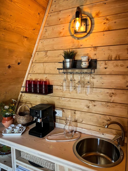 Wooden A-Frame cabin kitchenette with coffee machine, sink, wall shelves, glassware, and warm rope lighting at Guarda Che