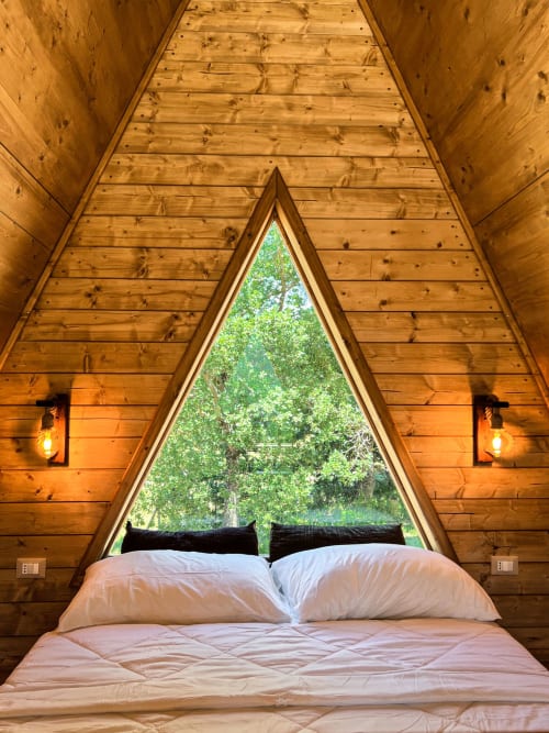 Cozy A-frame glamping bedroom with white bedding, mountain views through triangular window, warm wooden interior at Guarda