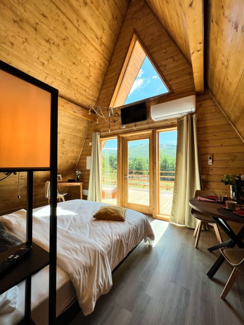 A-Frame glamping bedroom with wooden interior, mountain views through glass doors, bed, desk, and flat-screen TV at Guarda