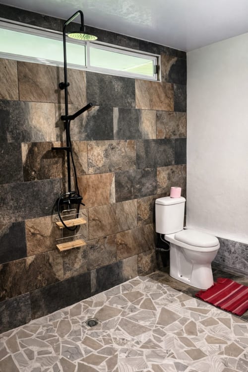 Salle de bain moderne à la Villa Vaipipiha à Paopao avec douche, robinetterie noire, murs en pierre et sol en mosaïque blanche.