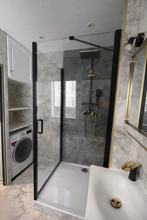 Salle de bains moderne avec douche à paroi vitrée noire, carrelage marbre, vasque blanche et robinetterie dorée à ATHEA
