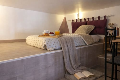 Chambre cosy d'appartement parisien avec lit surélevé, parure à pois, plateau petit-déjeuner, tête de lit bordeaux et éclairage chaleureux