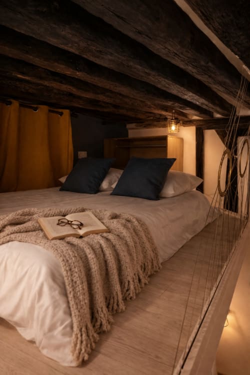 Chambre douillette sous les toits à l'appartement ATHEA Paris avec poutres apparentes, lit aux coussins noirs, couverture beige et coin lecture