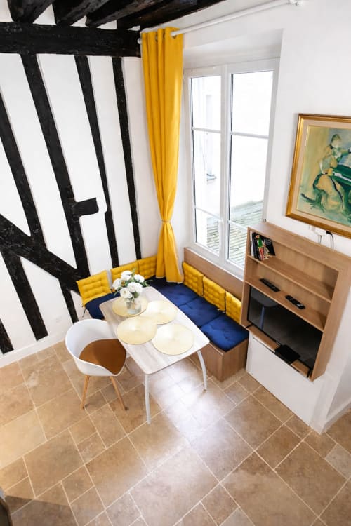 Salle à manger d'un appartement parisien moderne avec poutres noires, rideaux jaunes, banquette bleue et mobilier en bois