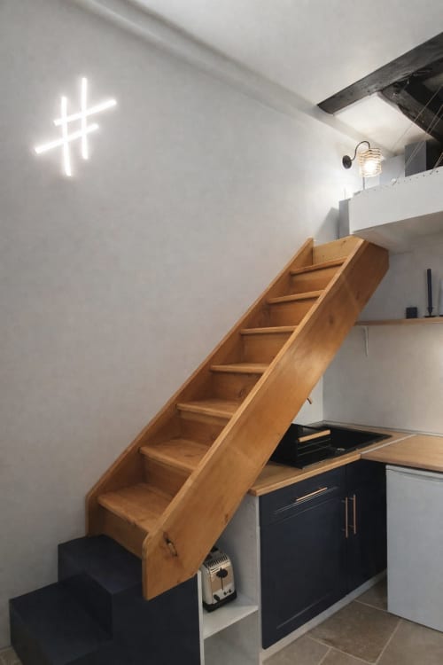 Loft moderne à ATHEA Paris avec escalier en bois, croix néon, kitchenette noire et murs blancs