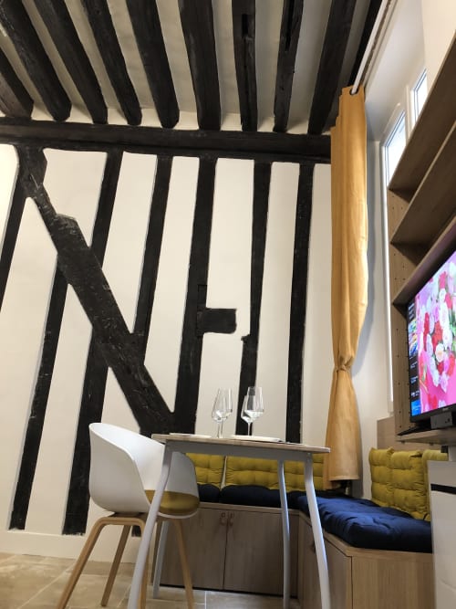 Intérieur d'appartement parisien moderne à ATHEA bnb avec poutre en bois noir, chaise blanche et lit aux oreillers jaunes