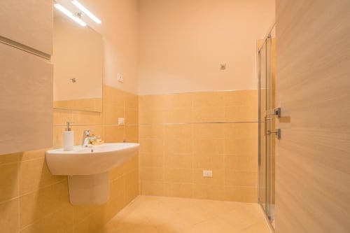 Bagno moderno con rivestimenti in piastrelle dorate, lavabo a colonna bianco e doccia con divisorio in vetro presso Le Alpi bed & living a Cividale
