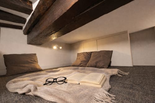 Chambre cosy du loft à l'appartement ATHEA Paris, poutres en bois apparentes, livre et lunettes sur le lit, coussins marron