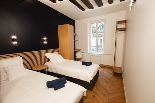 Chambre moderne de l'appartement ATHEA Paris avec deux lits, tête de lit rayée, mur d'accent noir, poutres bois et vue parisienne
