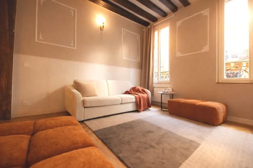 Salon lumineux d'appartement parisien avec canapé crème, poufs orange, poutres en bois et vue sur la ville par grandes fenêtres