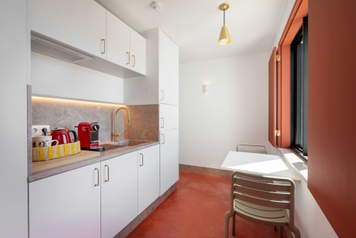 Modern kitchenette with white cabinetry, red kettle, coffee machine, and dining area in Monte das Açoteias studio apartment