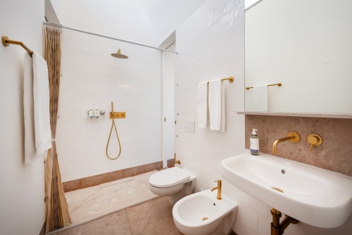 Modern bathroom with white walls, brown tile accents, brass fixtures, glass shower enclosure, and dual vanity sinks at Monte