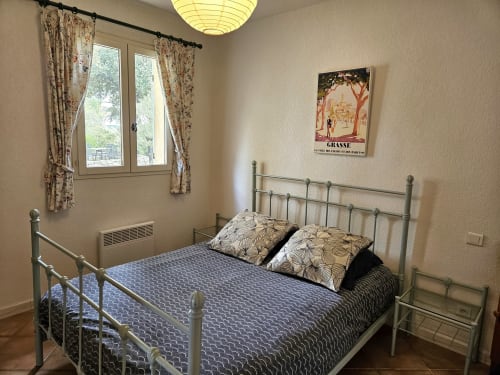 Chambre avec lit en métal, literie à motifs bleus, rideaux fleuris et affiche vintage de Grasse à la Villa Les Cyprès
