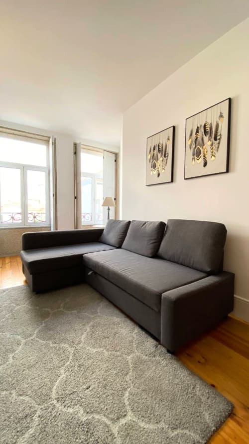 Sala moderna com sofá cinza escuro em L, paredes bege, quadros botânicos emoldurados, pisos de madeira e grandes janelas