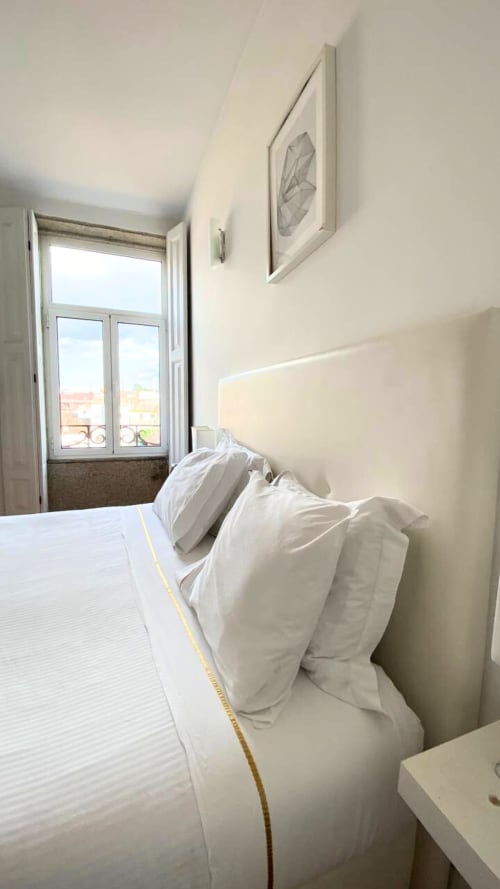 Quarto luminoso em apartamento no Porto com roupa de cama branca, vista da cidade pelas portas da varanda e quadros nas paredes bege