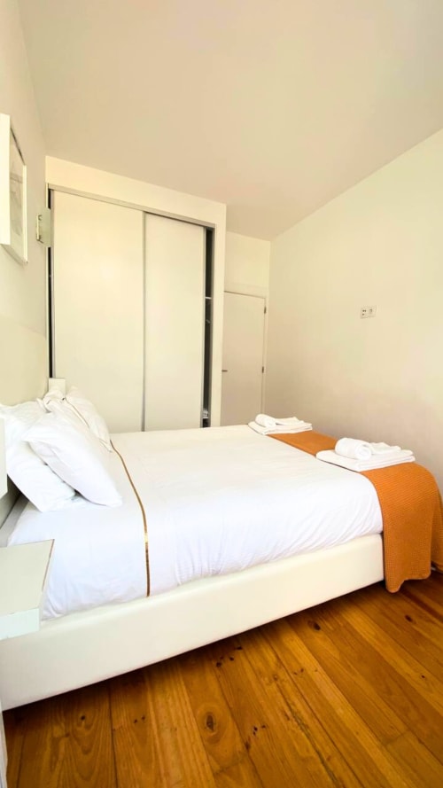 Quarto moderno no Bom Porto Apartments no Porto com cama branca, manta laranja, piso de madeira e portas de guarda-roupa deslizantes