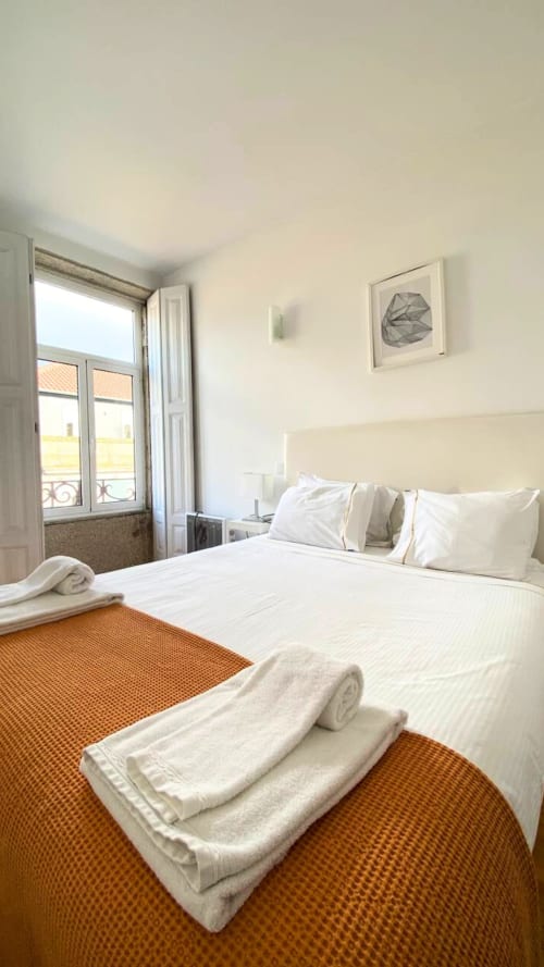 Quarto luminoso em apartamento do Porto com cama branca, tapete laranja e portas francesas com vista para a cidade