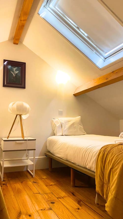 Aconchego do quarto na mansarda do apartamento no Porto com claraboia, vigas de madeira, cama, luminária tripé e piso em madeira