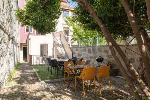 Pátio exterior do Bom Porto Apartments com mesas de refeição, cadeiras amarelas, árvores e muros de pedra no Porto