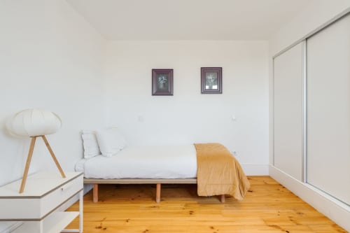 Quarto moderno no Bom Porto Apartments, Porto, com piso de madeira, cama branca com manta laranja, luminária pendente e vista para o jardim.
